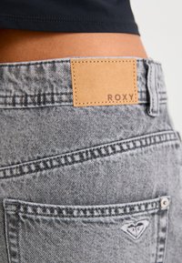 Pilki džinsai su odiniu užrašu "ROXY", išsiskiriantys siuvinėjimais, širdies emblemomis ir tekstūruotu paviršiumi.