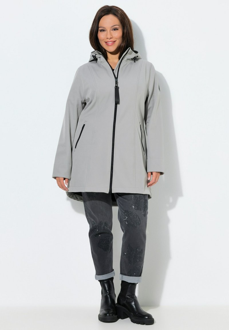 Žena v světle šedém kabátu s kapucí a černým zipem, tmavě šedých kalhotách s květinovými detaily a černých kotníkových botách, usmívající se, stojící.