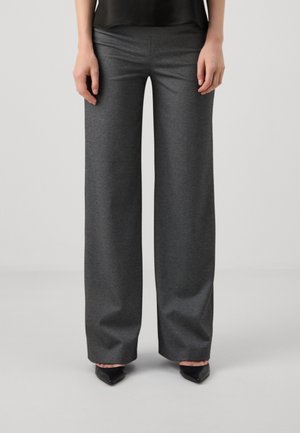 Vrouw draagt donkergrijze broek met wijde pijpen en zwarte schoenen met puntige neus, staand tegen een effen lichtgrijze achtergrond.