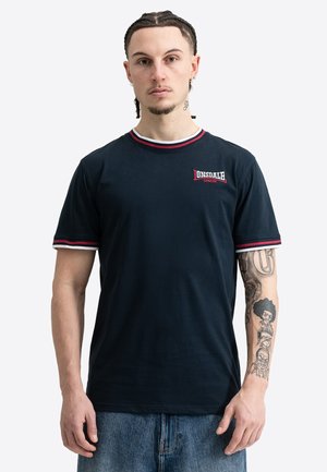 Giovane uomo con capelli intrecciati che indossa una maglietta nera Lonsdale London mostrando il braccio destro tatuato e jeans blu su sfondo bianco.