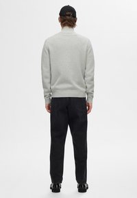 Grå tröja med hög krage, ribbat mönster; kombinerad med svarta jeansbyxor. Svart keps och kraftiga svarta skor kompletterar outfiten.