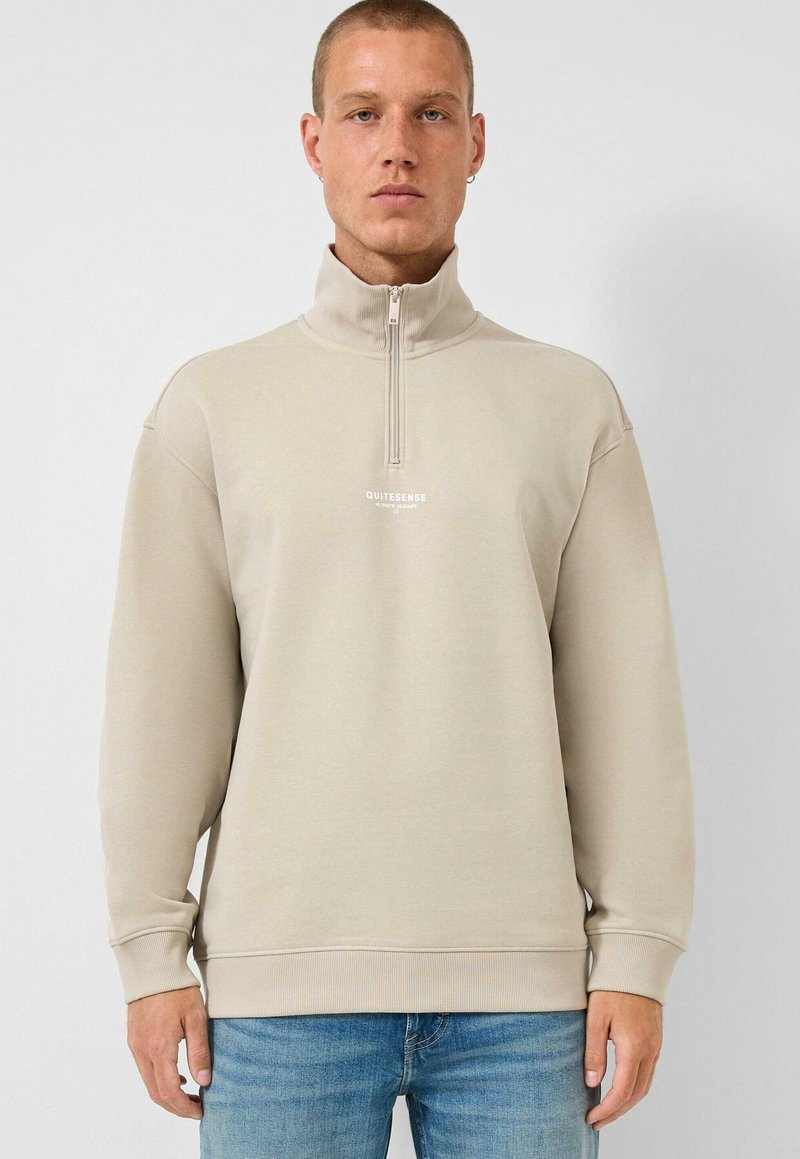Muž oblečený v béžové mikině s půlzipem a nápisem „QUITESENSE“, v kombinaci s modrými džíny, stojící před jednobarevným světlým pozadím.