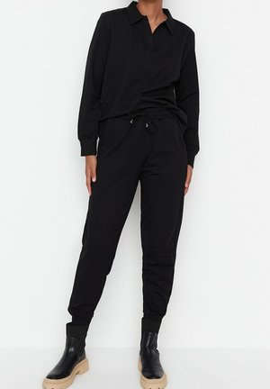 Person trägt ein schwarzes, langärmeliges Oberteil mit Kragen und schwarze Jogginghosen, kombiniert mit schwarzen Stiefeletten mit beigen Sohlen, steht vor einem weißen Hintergrund.