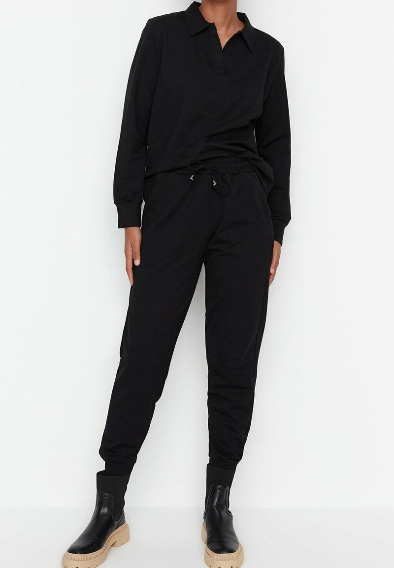 Personne portant un haut noir à manches longues avec col et un pantalon de jogging noir, assortis à des bottines noires à semelles beige, debout devant un fond blanc.