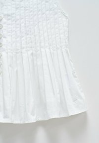 Top blanco sin mangas con pliegues verticales, que presenta un cierre frontal con botones y un ligero vuelo en el dobladillo. Textura de algodón suave.