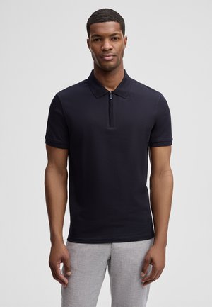 Mann trägt ein schwarzes Poloshirt mit kurzen Ärmeln und Frontreißverschluss sowie hellgraue Hose, steht vor schlichtem Hintergrund.