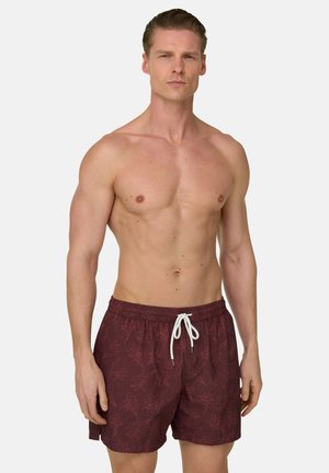 Uomo senza maglietta con capelli corti che indossa pantaloncini da bagno color bordeaux con motivo floreale e coulisse bianca, in piedi davanti a uno sfondo semplice.