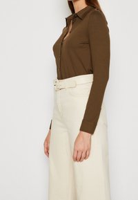 Top marrón de manga larga con cuello y botones delanteros. Combinado con pantalones blancos de talle alto y pierna ancha, y un cinturón beige. Textura lisa.