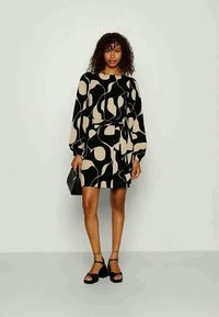 Črna in bež obleka s potiskom, dolgi rokavi, zategnjen pas in razširjen rob. Obuta v črne čevlje in z majhno torbico.