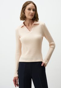 Perzikkleurige gebreide pullover met lange mouwen en een V-hals, voorzien van een contrasterende kraag. De geweven stof heeft een geruit patroon en wordt gecombineerd met donkere broeken.