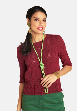 Woman wearing a maroon knit top, green beaded necklace, flower-shaped earrings, green pants, and a gold bracelet against a white background.