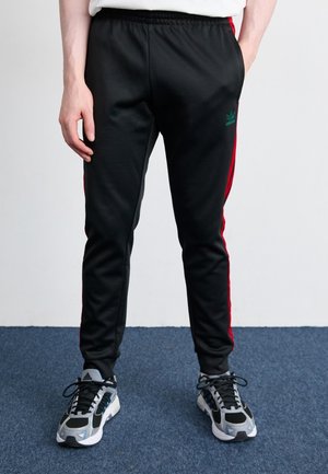 Person iført sorte Adidas træningsbukser med røde striber og grå Adidas sneakers står på blåt gulvtæppe.