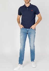 Camisa polo azul marino con collar y mangas cortas, combinada con jeans claros desgastados y zapatillas blancas, mostrando un estilo casual.