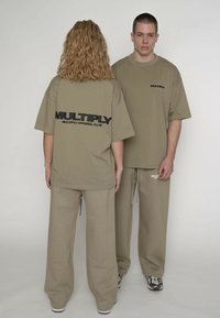 Oversized t-shirts og matchende bukser i khaki med sort tekst på ryggen. Stoffet virker blødt med en afslappet pasform. Detaljer inkluderer en snor til justering.
