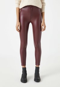 Kvinna iklädd högmidjade bordeauxfärgade imitationläderleggings och svarta ankelskor, kombinerat med en beigemelerad stickad tröja.