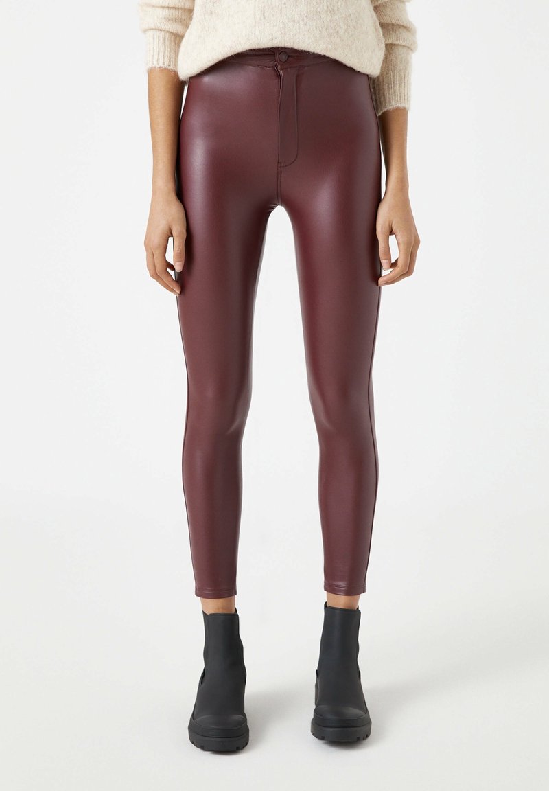 Kvinna iklädd högmidjade bordeauxfärgade imitationläderleggings och svarta ankelskor, kombinerat med en beigemelerad stickad tröja.