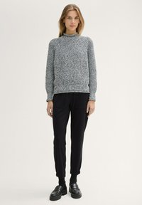Grauer Strickpullover mit hohem Kragen, kombiniert mit schwarzen Jogginghosen. Das Outfit hat gerippte Bündchen und eine lockere Passform. Schwarze Slipper.
