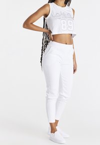 Weißes, kürz geschnittenes Sportoberteil mit dem Schriftzug "SikSilk 89", kombiniert mit weißen, enganliegenden Hosen, die zwei Fronttaschen und eine glatte Textur aufweisen.