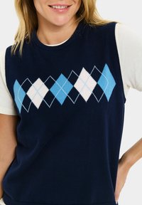 Hymyilevä henkilö, joka käyttää tummansinistä argyle-neuleliiviä, jossa on vaaleansinisiä ja valkoisia timanttiesityksiä valkoisen lyhytnuppuisen paidan päällä.