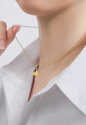 Mujer con un collar colgante de forma de abanico dorado, sosteniendo la cadena cerca del cuello, vestida con una camisa blanca con el cuello abierto.