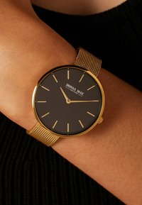 Horloge met goudkleurige kast en zwarte wijzerplaat, minimalistische uurmarkeringen en een meshband. Merkwaarde "DONNA MAE" zichtbaar op de wijzerplaat.