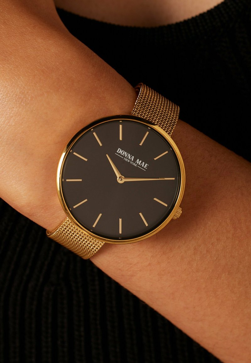 Horloge met goudkleurige kast en zwarte wijzerplaat, minimalistische uurmarkeringen en een meshband. Merkwaarde "DONNA MAE" zichtbaar op de wijzerplaat.