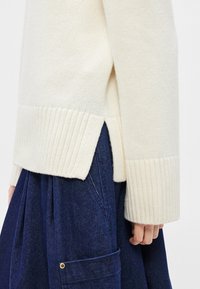 Πουλόβερ πλεκτό κρεμ με ραβδώσεις στα μανίκια και το τελείωμα, με πλευρικές σχισμές. Συνδυάζεται με σκουρόχρωμη φούστα από τζιν.