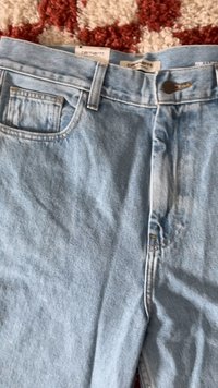 Lichtblauwe denim jeans met metalen knoop en Carhartt merklabels op een rood-witte getextureerde ondergrond.