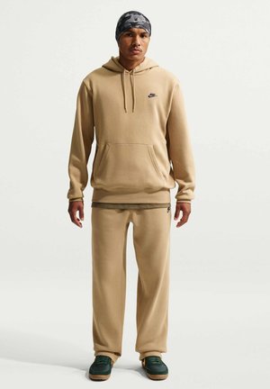 Mężczyzna w beżowej bluzie z kapturem i spodniach dresowych Nike, zielonych butach sportowych i szarej czapce moro, stojący na jednolitym tle.