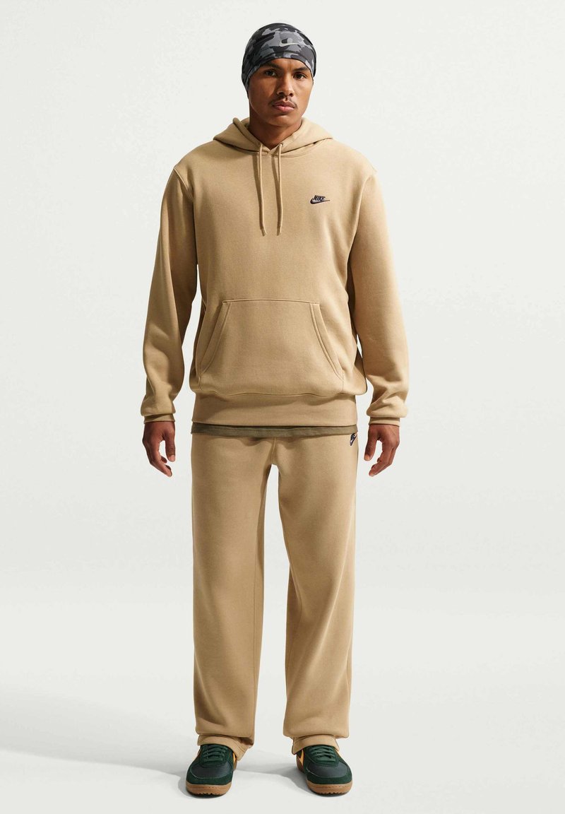 Mann trägt beige Nike-Hoodie und Jogginghose, grüne Sneakers und eine graue Camouflage-Mütze, steht vor schlichtem Hintergrund.