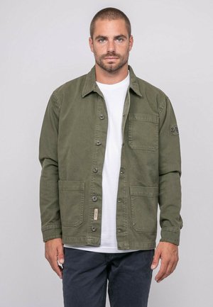 Homme aux cheveux courts et à la barbe, portant une veste boutonnée vert olive sur un t-shirt blanc et un pantalon foncé, debout devant un fond uni.