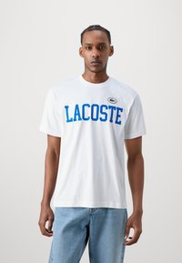 Balta medvilnės marškinėliai su mėlynu "LACOSTE" užrašu, trumpomis rankovėmis, apvaliu iškirpimu ir mažuoju logotipo pleistru viršutiniame kairiajame šone. Dėvimi su šviesiai mėlynomis džinsais.
