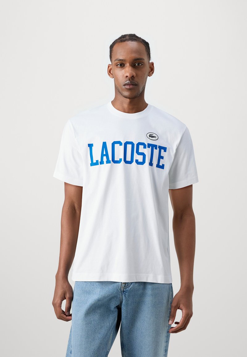 Balta medvilnės marškinėliai su mėlynu "LACOSTE" užrašu, trumpomis rankovėmis, apvaliu iškirpimu ir mažuoju logotipo pleistru viršutiniame kairiajame šone. Dėvimi su šviesiai mėlynomis džinsais.