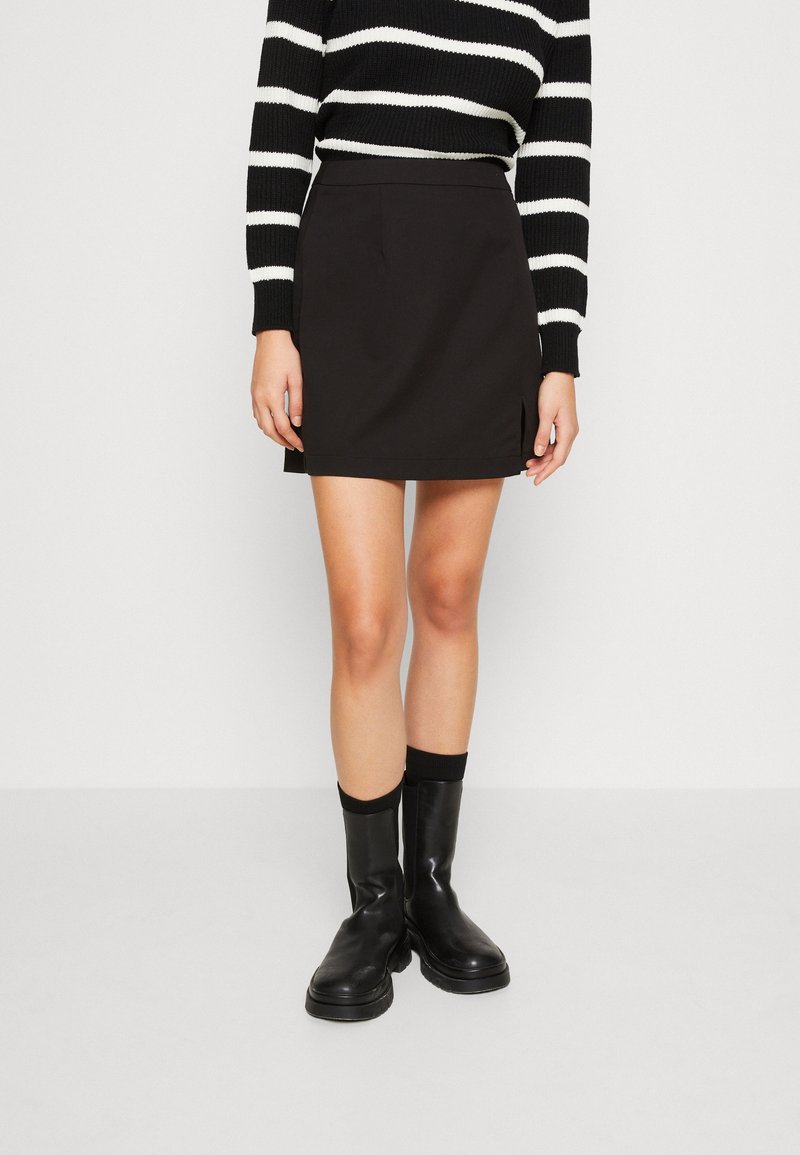 Femme portant une mini-jupe noire, un haut à manches longues rayé noir et blanc, et des bottines noires, debout devant un fond uni.