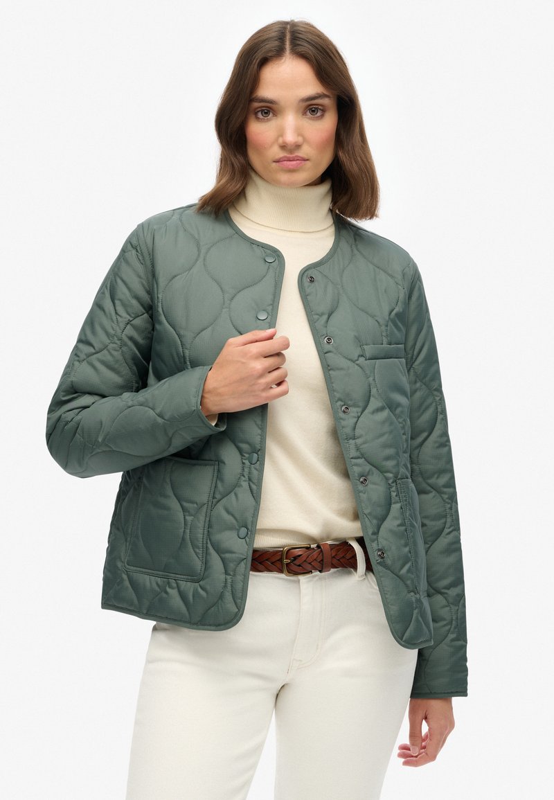 Gequilted groene jas met een ronde halslijn, drukknopen en een voorkoop. Draag het over een beige coltrui en crèmekleurige broek.