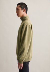 Jongeman met kort zwart haar, gekleed in een olijfgroene zip-up sweatshirt en zwarte broek, staand in profiel tegen een eenvoudige achtergrond.