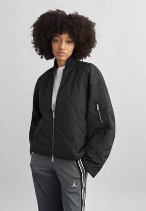 Fiatal nő göndör hajjal, fekete steppelt dzsekit és szürke sportnadrágot visel fehér csíkokkal és logóval, egyszínű háttér előtt áll.