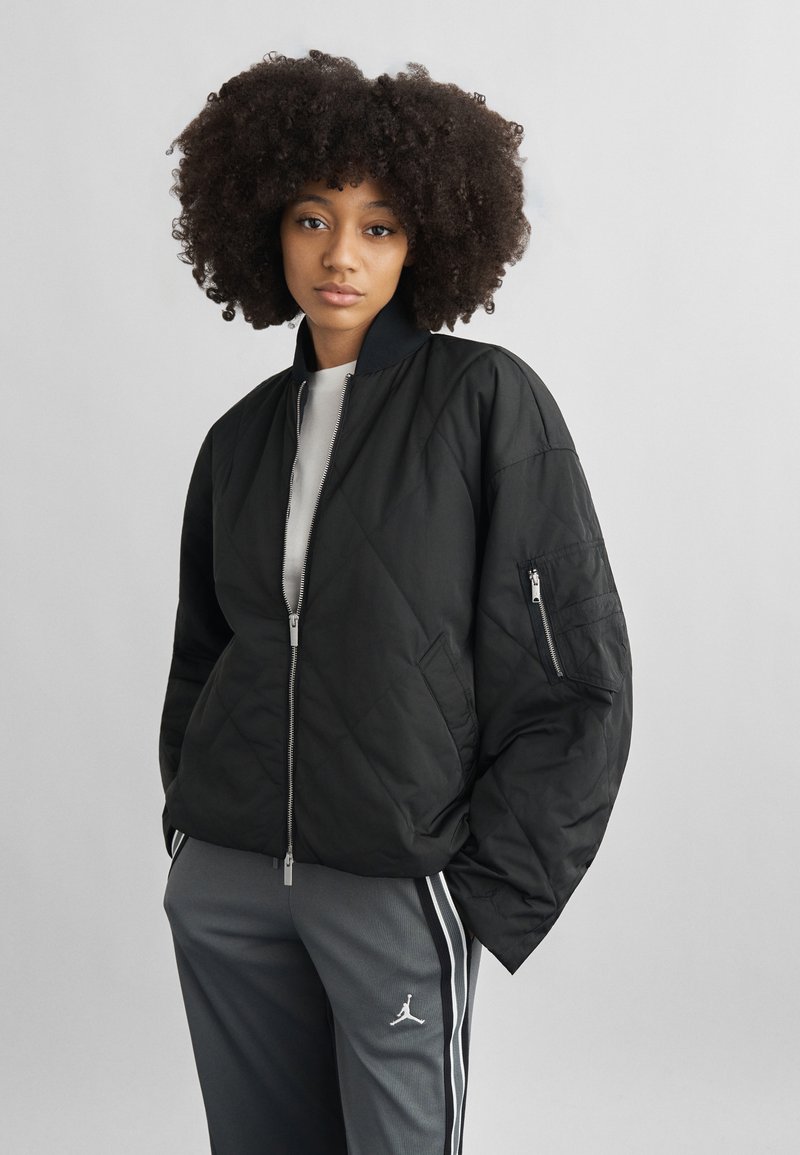 Νεαρή γυναίκα με σγουρά μαλλιά, φορώντας μαύρο μπουφάν με ραφές και γκρι αθλητικό παντελόνι με λευκές ρίγες και λογότυπο, στέκεται μπροστά σε απλό φόντο.