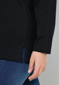 Schwarzes Oberteil mit langen Ärmeln und seitlichen Schlitzen, aus weichem Stoff. Kombiniert mit blauen Jeans, die einen Nahaufnahme des Saums und der Hand zeigen.