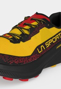 Κίτρινο, κόκκινο και μαύρο παπούτσι trail running La Sportiva με μαύρα κορδόνια και ανθεκτική μαύρη σόλα με κόκκινες λεπτομέρειες πέλματος.
