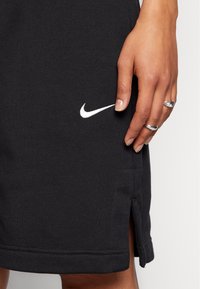 Närbild av en hand som bär silverringar, vilande på ett svart Nike-plagg med en vit swoosh-logotyp och sidopenna.