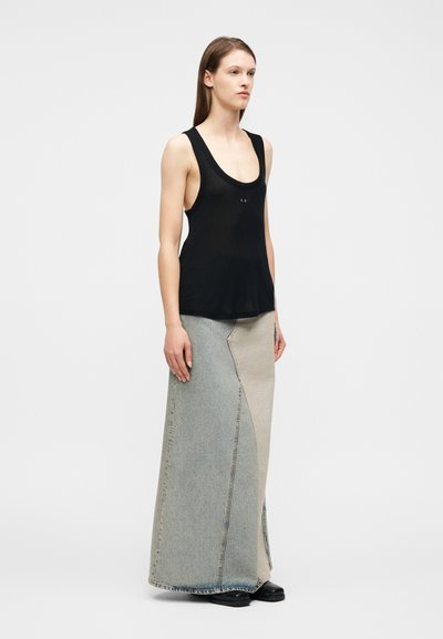 Γυναίκα που φοράει μαύρο τοπ χωρίς μανίκια και μακριά φούστα από τζιν σε δύο αποχρώσεις, στέκεται μπροστά σε απλό ανοιχτό φόντο.