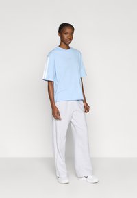 Világoskék pamut rövid ujjú póló fehér díszcsíkkal és kis logóval. Szürke melegítőnadrággal és fehér cipővel párosítva.