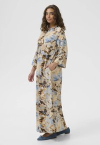 Pitkä, luidenkaltainen mekko vaaleanruskeana, jossa on sinisiä ja ruskeita abstrakteja kuvioita. Siinä on pyöreä pääntie, pitkät hihat ja sivutaskut. Yhdistetty sinisiin ballerinoihin.