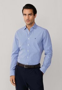 Muškarac nosi plavu košulju na kockice s kopčanjem, uvučenu u crne hlače s smeđim remenom, stoji s jednom rukom u džepu.