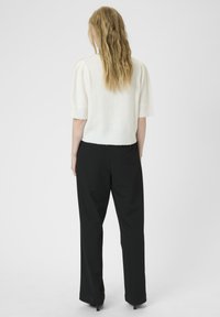 Vrouw met lang golvend blond haar, gekleed in een wit kort mouwtopje en zwarte broek met wijde pijpen, staand met de rug naar de camera op een effen achtergrond.