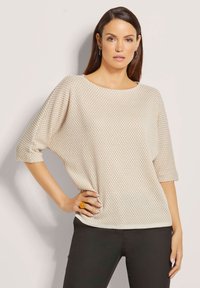 Beige gebreide trui met een gewatteerd honingraatpatroon, voorzien van een brede halslijn en driekwartmouwen, gecombineerd met zwarte broek.