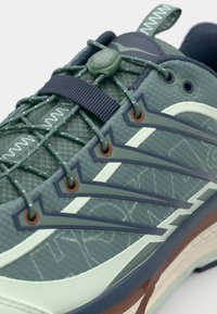 Zielone buty sportowe z teksturowanym wierzchem, kontrastującymi granatowymi akcentami, elastycznymi sznurowadłami oraz plastikowym zapięciem do regulacji.