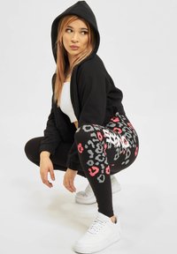 Svart hettejakke med glidelås, svarte leggings med rød og grå leopardmønster, og hvite joggesko. Modellen sitter i en avslappet posisjon.