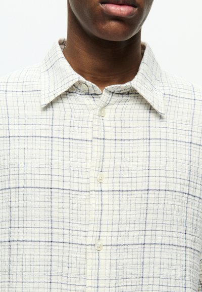 Chemise boutonnée couleur crème clair avec un motif à carreaux bleus. Présente un col et de petits boutons blancs, fabriquée en tissu texturé.